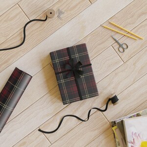 K&ouml;nnte beinhalten: Ein verpacktes Geschenk mit schwarzer Schleife, schwarzem Band und Geschenkpapier mit einem rotbraunen, schwarzen und goldenen Karomuster. Au&szlig;erdem eine Rolle Geschenkpapier, eine Schere und Bleistifte auf einer hellen Holzoberfl&auml;che.