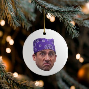 May include: A white, round Christmas ornament featuring a close-up of a person's face wearing a purple bandana. The person has a concerned expression. The ornament hangs from a gold string against a blurred background of Christmas lights and greenery.