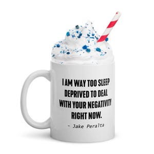 May include: White ceramic mug with a black quote that reads "I am way too sleep deprived to deal with your negativity right now. - Jake Peralta". The mug has a white rim and handle. The mug is filled with white whipped cream and blue sprinkles. A red and white striped straw is in the mug.