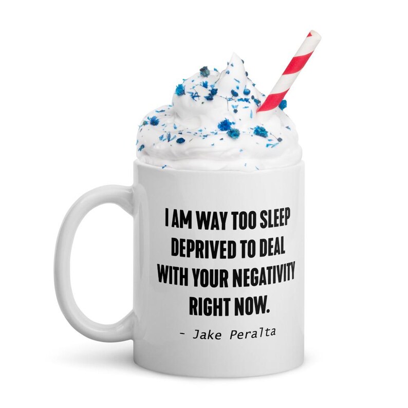 May include: White ceramic mug with a black quote that reads "I am way too sleep deprived to deal with your negativity right now. - Jake Peralta". The mug has a white rim and handle. The mug is filled with white whipped cream and blue sprinkles. A red and white striped straw is in the mug.