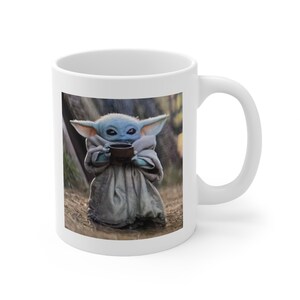 Puede incluir: Taza de cer&aacute;mica blanca con una imagen colorida de un peque&ntilde;o personaje ficticio verde sosteniendo una taza. El personaje tiene orejas grandes y ojos grandes. El fondo es una imagen borrosa de un bosque.