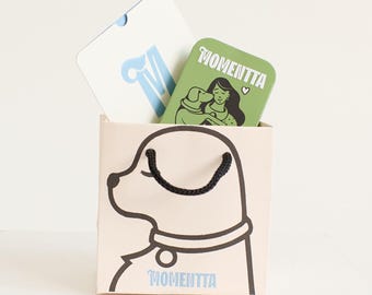 Puede incluir: Una bolsa de regalo beige con una ilustración de perro negro y la palabra "MOMENTTA" impresa. Dentro hay dos tarjetas: una blanca con una "M" azul y otra verde con una mujer y la palabra "MOMENTTA".