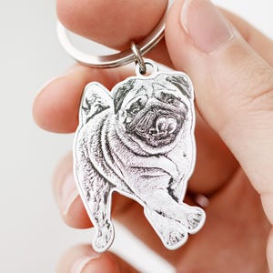 Puede incluir: Un llavero plateado con un grabado detallado de un perro pug. El pug se representa en una pose de pie con la lengua ligeramente fuera. El llavero tiene un anillo circular para sujetar las llaves.