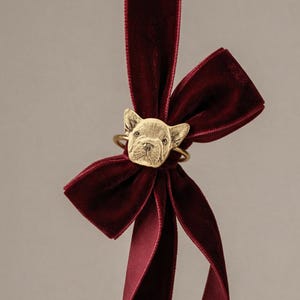 Puede incluir: Un anillo dorado con un dije detallado en forma de cabeza de bulldog francés, centrado en una cinta de terciopelo color burdeos atada en forma de lazo. El dije de metal muestra las facciones del perro, incluyendo orejas puntiagudas.