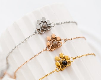 May include: Three delicate bracelets with paw print charms. The bracelets are in silver, rose gold, and gold tones. Each charm has a small, round center. The bracelets are displayed on a white, textured surface.