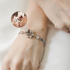 May include: A silver bracelet featuring a heart-shaped charm with a paw print design. The bracelet has clear, round accent stones and a small blue enamel detail. A small photo of a white dog is in the background.