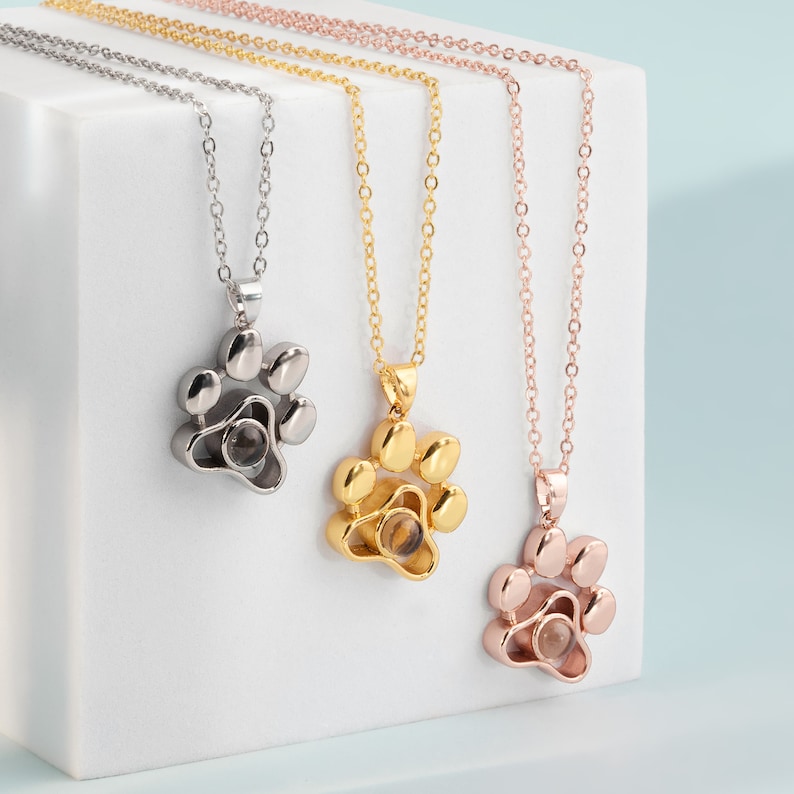 May include: Three necklaces with paw print pendants in silver, gold, and rose gold. Each pendant features a small, clear, round stone. The chains are delicate, and the pendants are three-dimensional.