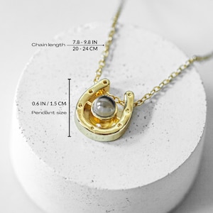 May include: A gold-colored horseshoe pendant necklace with a chain length of 7.8 to 9.8 inches (20-24 cm). The pendant is 0.6 inches (1.5 cm) in size and features a round, gray stone in the center. The necklace is displayed on a white, textured surface.