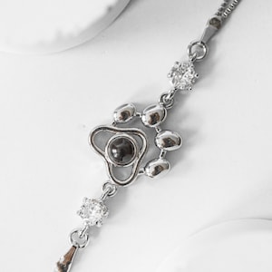 May include: A silver-toned bracelet featuring a paw print design with a dark center stone and clear, round accent stones. The bracelet has a delicate chain and a small, teardrop-shaped charm at the end.