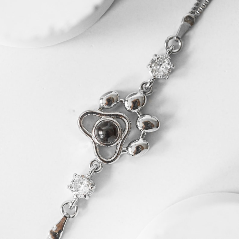 May include: A silver-toned bracelet featuring a paw print design with a dark center stone and clear, round accent stones. The bracelet has a delicate chain and a small, teardrop-shaped charm at the end.