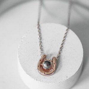 May include: A rose gold horseshoe pendant necklace with a silver chain. The horseshoe has a small, round, gray stone in the center. The necklace is displayed on a white, circular surface.