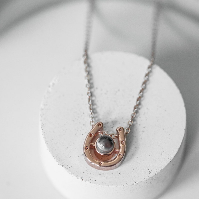 May include: A rose gold horseshoe pendant necklace with a silver chain. The horseshoe has a small, round, gray stone in the center. The necklace is displayed on a white, circular surface.