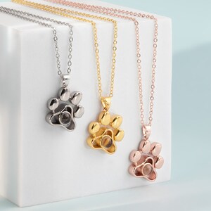 May include: Three necklaces with paw print pendants. The pendants are silver, gold, and rose gold. Each paw print pendant has a small, round center. The necklaces are displayed on a white surface against a light blue background.