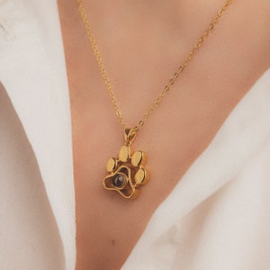 May include: Gold-coloured paw print pendant necklace. The pendant features a detailed paw print design with a small, dark centre. The necklace is on a delicate gold chain.