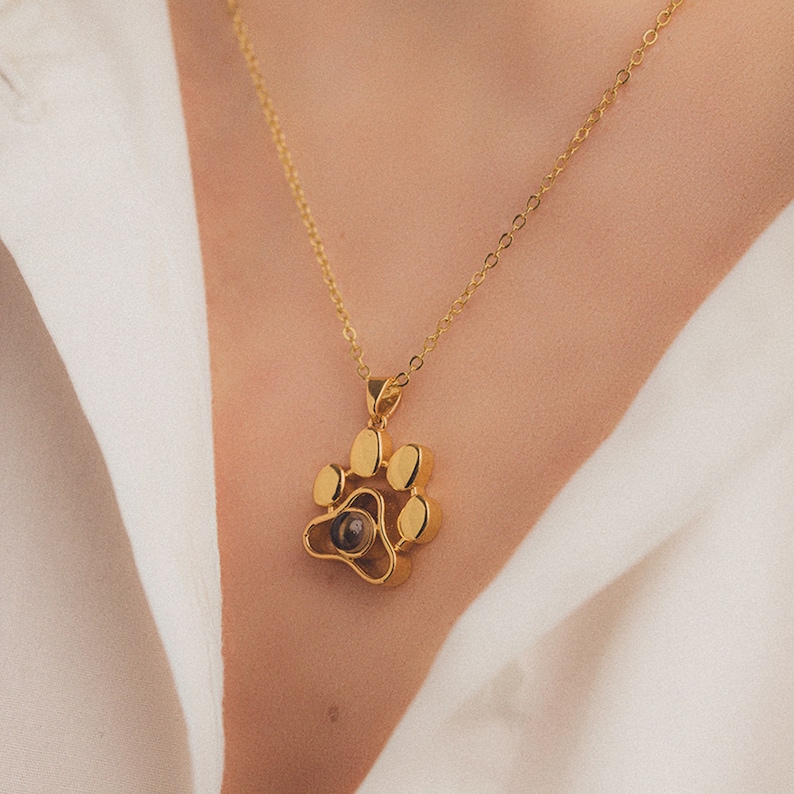 May include: Gold-coloured paw print pendant necklace. The pendant features a detailed paw print design with a small, dark centre. The necklace is on a delicate gold chain.