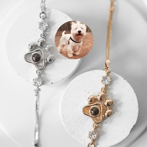 May include: Two necklaces with paw print pendants. One is silver, the other gold. Each pendant has a central dark stone and is accented with clear crystals. A small photo of a white dog is in the background.