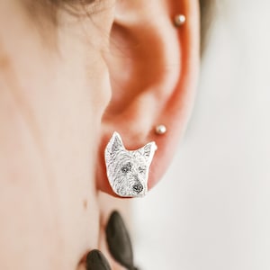 Puede incluir: Pendientes de botón plateados con forma de cabeza de perro, con detalles finos. Los pendientes se lucen en una oreja, destacando el diseño y la artesanía. Un accesorio único para los amantes de los perros.
