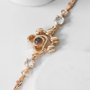 May include: Gold-toned bracelet featuring a paw print design with a central brown stone and clear crystals. The bracelet has a delicate chain and is set against a white background.