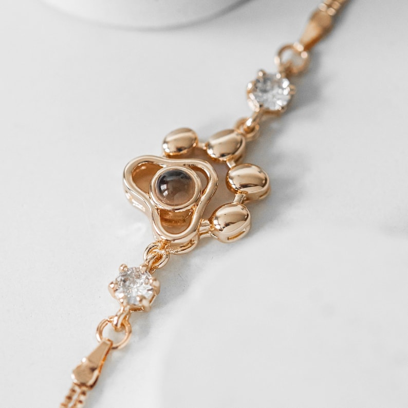 May include: Gold-toned bracelet featuring a paw print design with a central brown stone and clear crystals. The bracelet has a delicate chain and is set against a white background.