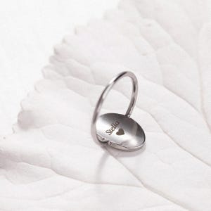 Puede incluir: Un anillo plateado con una cara circular y una banda curva. La cara del anillo tiene grabado el nombre "Stella", junto con un peque&ntilde;o s&iacute;mbolo de coraz&oacute;n. El anillo est&aacute; sobre un fondo blanco.