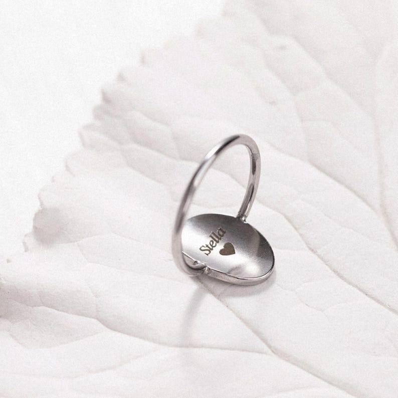 Puede incluir: Un anillo plateado con una cara circular y una banda curva. La cara del anillo tiene grabado el nombre "Stella", junto con un peque&ntilde;o s&iacute;mbolo de coraz&oacute;n. El anillo est&aacute; sobre un fondo blanco.