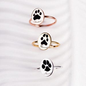 Puede incluir: Tres anillos con caras ovaladas, cada uno con una huella de pata negra y un nombre en cursiva. Los anillos son de oro rosa, oro y plata. El fondo es una superficie blanca texturizada.