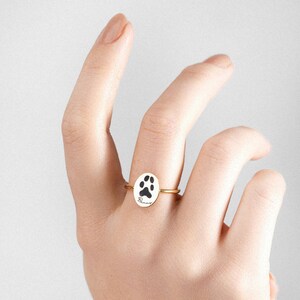 Puede incluir: Anillo dorado con un colgante ovalado que presenta una huella de pata negra y la palabra "Bunny" en cursiva. El anillo se muestra sobre un fondo neutro, resaltando el dise&ntilde;o y los detalles de la joya. Una delicada pieza de joyer&iacute;a con tem&aacute;tica de mascotas.