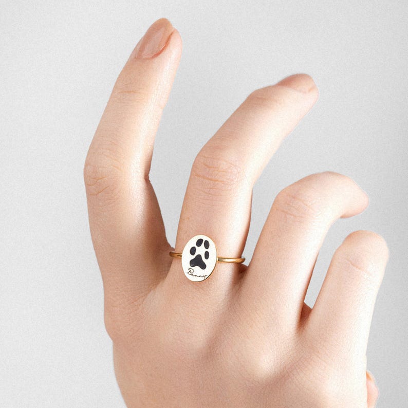 Puede incluir: Anillo dorado con un colgante ovalado que presenta una huella de pata negra y la palabra "Bunny" en cursiva. El anillo se muestra sobre un fondo neutro, resaltando el dise&ntilde;o y los detalles de la joya. Una delicada pieza de joyer&iacute;a con tem&aacute;tica de mascotas.