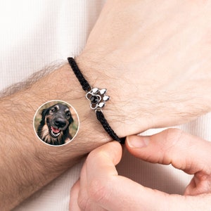 Könnte beinhalten: Schwarz geflochtenes Armband mit einem silbernen Pfotenabdruck-Charm mit einem schwarzen Mittelstein. Das Armband wird am Handgelenk getragen. Ein kleines rundes Bild eines Hundes befindet sich in der unteren linken Ecke.
