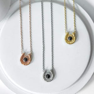 May include: Three horseshoe pendant necklaces in rose gold, silver, and gold. Each necklace features a small, round, black stone set in the center of the horseshoe. The chains are delicate and the pendants are displayed on a white surface.