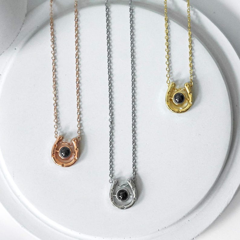 May include: Three horseshoe pendant necklaces in rose gold, silver, and gold. Each necklace features a small, round, black stone set in the center of the horseshoe. The chains are delicate and the pendants are displayed on a white surface.