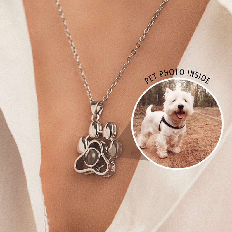 May include: A silver paw print pendant necklace with a chain. The pendant has a detailed paw print design and a circular inset. The image includes a white dog with the text "PET PHOTO INSIDE".