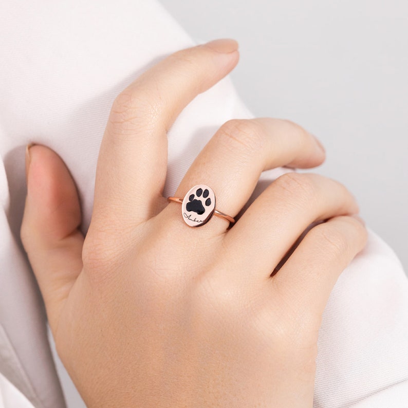 Puede incluir: Anillo de oro rosa con un colgante ovalado con una huella de pata negra. El anillo tiene una banda delgada y el nombre "Amber" est&aacute; escrito en cursiva. El anillo se muestra en el dedo de una persona.