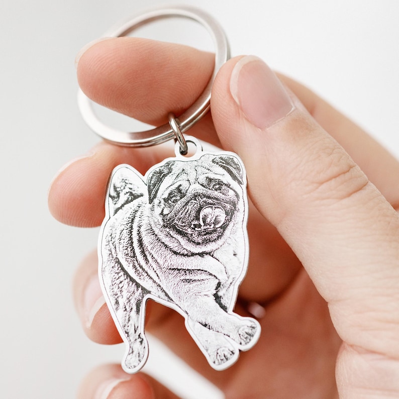 Puede incluir: Un llavero plateado con un grabado detallado de un perro pug. El pug se representa en una pose de pie con la lengua ligeramente fuera. El llavero tiene un anillo circular para sujetar las llaves.