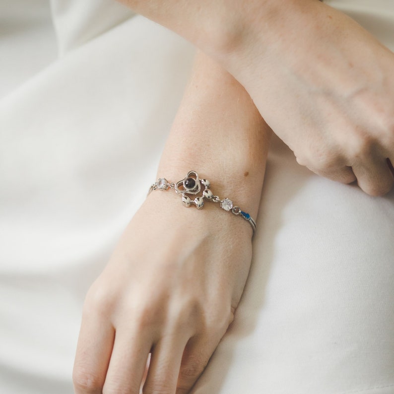 May include: A silver bracelet with a floral design and a central black stone. The bracelet features clear and blue accent stones. The bracelet is displayed on a person's wrist.