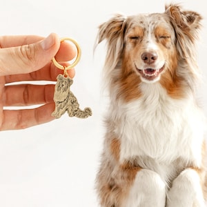 Puede incluir: Un llavero de metal dorado con una silueta detallada de un perro. El perro tiene un pelaje esponjoso y est&aacute; sentado. El llavero se sostiene frente a un perro feliz y sonriente con pelaje marr&oacute;n, blanco y canela.