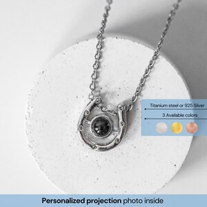 May include: A silver horseshoe pendant necklace with a chain. The pendant has a dark center stone and is on a white surface. Text on the image says "Titanium steel or 925 Silver" and "3 Available colors". The text "Personalized projection photo inside" is also present.