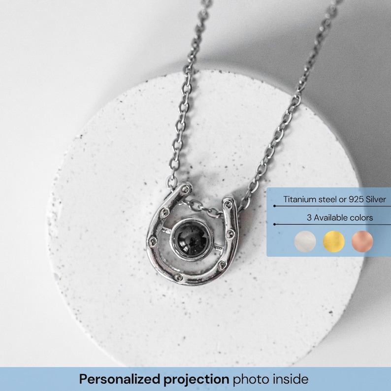 May include: A silver horseshoe pendant necklace with a chain. The pendant has a dark center stone and is on a white surface. Text on the image says "Titanium steel or 925 Silver" and "3 Available colors". The text "Personalized projection photo inside" is also present.