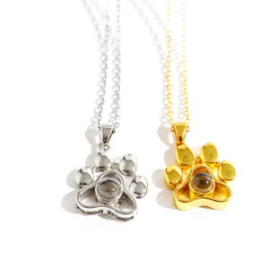 May include: Two necklaces with paw print pendants. One pendant is silver-colored, the other is gold-colored. Each pendant has a small, round, dark-colored stone in the center. The necklaces are on a white background.