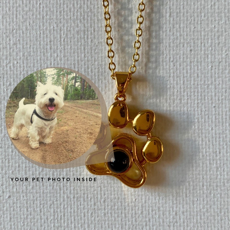 May include: Gold-coloured paw print pendant necklace with a chain. The pendant has a black stone in the centre and is next to a circular image of a white dog. The text "YOUR PET PHOTO INSIDE" is below the image.