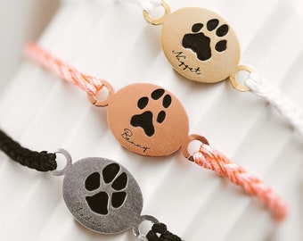 May include: Three personalized bracelets with paw print charms. The charms are in gold, copper, and silver tones, each with a black paw print and a name. The bracelets have white, coral, and black cords.