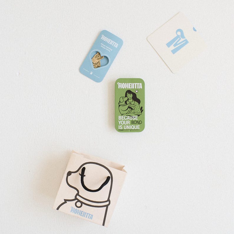 Puede incluir: Un surtido de art&iacute;culos de la marca Momentta sobre una superficie blanca. Incluye un im&aacute;n azul claro con forma de gato, un im&aacute;n rectangular verde con una ilustraci&oacute;n de mujer y perro, una peque&ntilde;a bolsa de papel con una ilustraci&oacute;n de perro y una tarjeta blanca.