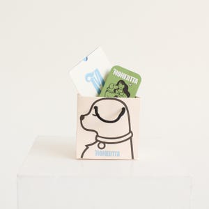 Puede incluir: Una bolsa de regalo beige con una ilustraci&oacute;n de perro negro y la palabra "MOMENTTA" impresa. Dos tarjetas dentro: una blanca con una "M" azul y otra verde con una mujer y la palabra "MOMENTTA".