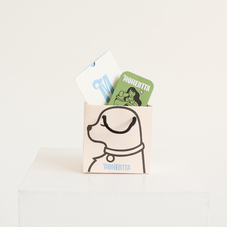 Puede incluir: Una bolsa de regalo beige con una ilustraci&oacute;n de perro negro y la palabra "MOMENTTA" impresa. Dos tarjetas dentro: una blanca con una "M" azul y otra verde con una mujer y la palabra "MOMENTTA".