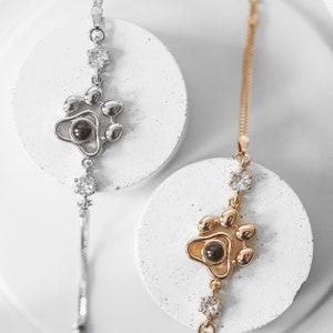 May include: Two necklaces with paw print pendants. One is silver-toned, the other gold-toned. Both feature a paw print design with a dark center and clear gemstone accents. The necklaces are displayed on white circular platforms against a white background.