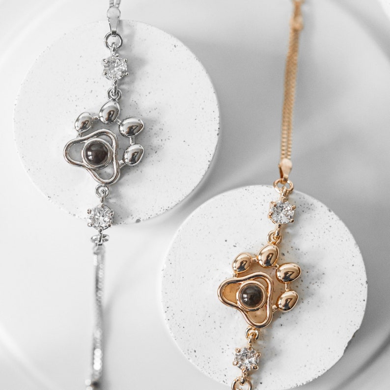 May include: Two necklaces with paw print pendants. One is silver-toned, the other gold-toned. Both feature a paw print design with a dark center and clear gemstone accents. The necklaces are displayed on white circular platforms against a white background.