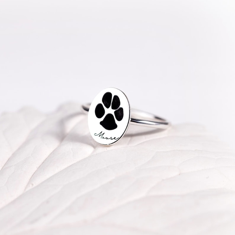 Puede incluir: Un anillo de plata con un colgante ovalado que presenta una huella de pata negra y el nombre "Moose" en escritura cursiva. El anillo se encuentra sobre un fondo blanco, resaltando su dise&ntilde;o y detalles.