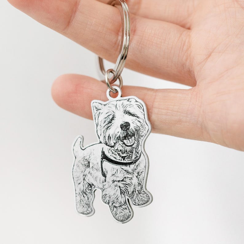Puede incluir: Un llavero plateado con una ilustraci&oacute;n detallada de un perro West Highland White Terrier. El perro est&aacute; representado en un estilo de boceto en blanco y negro, con un collar. El llavero est&aacute; unido a un anillo plateado.