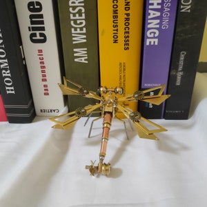 May include: A gold metal dragonfly sculpture with intricate details and gears. The dragonfly is perched on a stand with a copper-colored body.