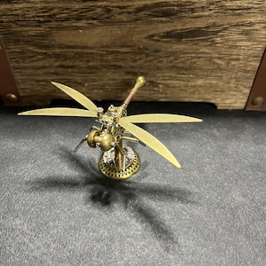 May include: A gold metal dragonfly sculpture with intricate details. The dragonfly has four wings and a long, thin body. The sculpture is mounted on a small, round base.